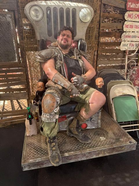 Brisket in spiked pauldron and rusted armor plates lounges on a throne built from a Jeep front grille, baby-doll heads mounted on either side.