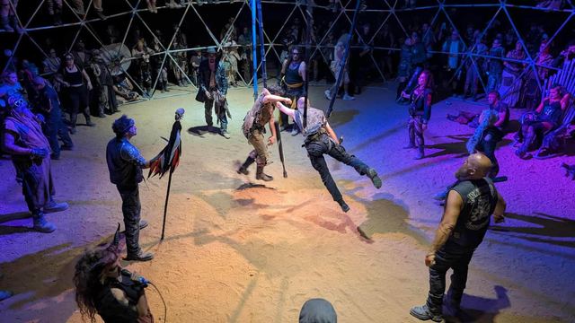 A fighter leaps mid-swing inside the thunderdome as the crowd watches from the lattice and the sand.