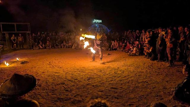 A fire performer in a horned mask breathes a jet of flame before a night crowd ringed around a sand arena, a geodesic dome glowing behind.