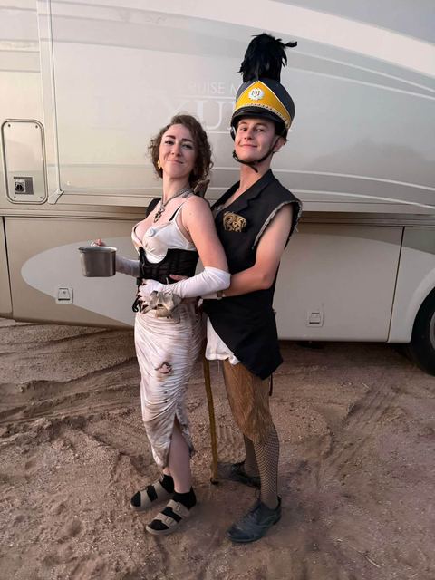Two wastelanders pose back to back in front of an RV — one in a torn white dress holding a tin cup, one in a feathered marching-band shako and cut-off blazer.