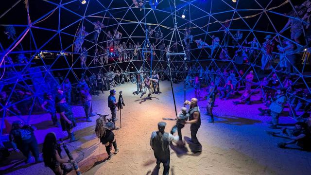 Inside a geodesic dome at night lit blue and orange, spectators climb the struts while fighters square off on the sand floor below.