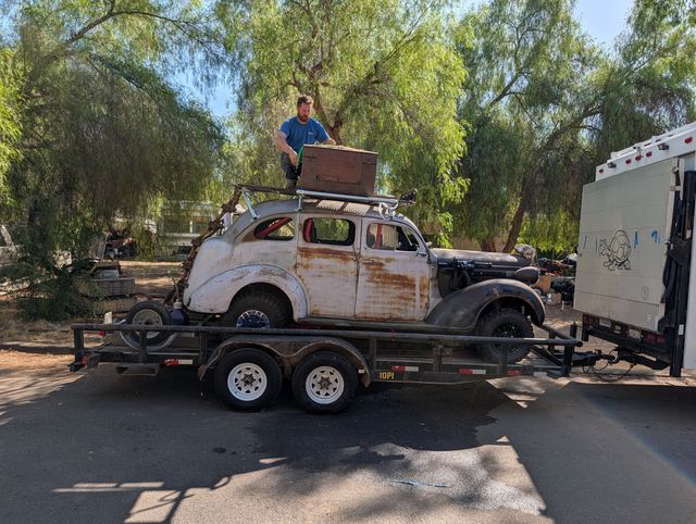A rusted 1930s sedan war car strapped to a flatbed trailer behind an RV on a tree-lined street, a crew member working on its roof.