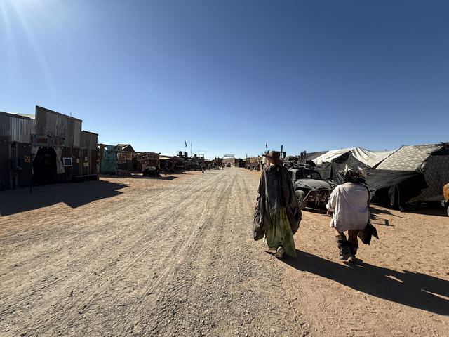 Wastelanders in long robes walking down the dusty main street of the festival town.