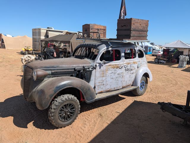 The war car parked at camp: rust-and-primer paint, bars over the windows, wooden crates on the roof.