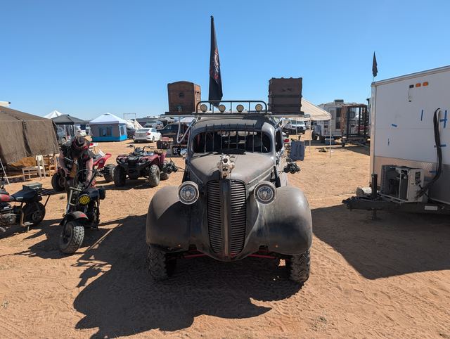 Head-on view of a black war car with a tall black flag, roof crates and spotlights, at camp.