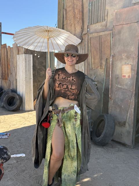 The same wastelander with the white parasol, posing in a long green skirt by the outpost door.