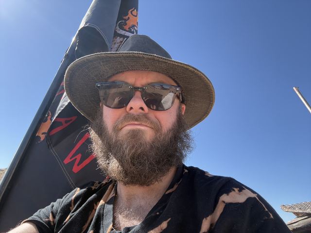 Selfie of a bearded man in a wide-brimmed hat and sunglasses against a blue sky.
