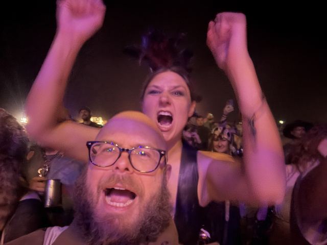 A bearded man in glasses and a woman with a feathered headdress yell with arms raised in a night crowd.