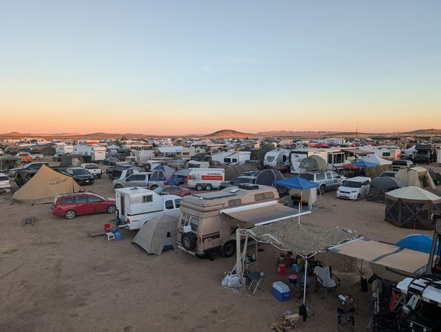 Rooftop panorama of the camp sea, canopies and campers under a pink sky.
