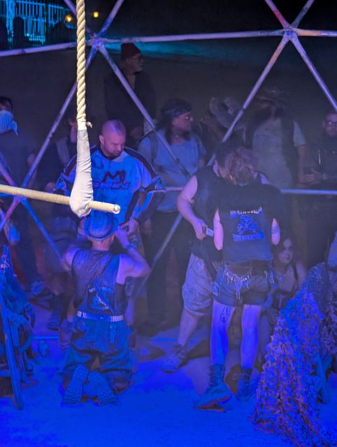 Inside the blue-lit Thunderdome, fighters and a referee gather beneath a hanging rope.