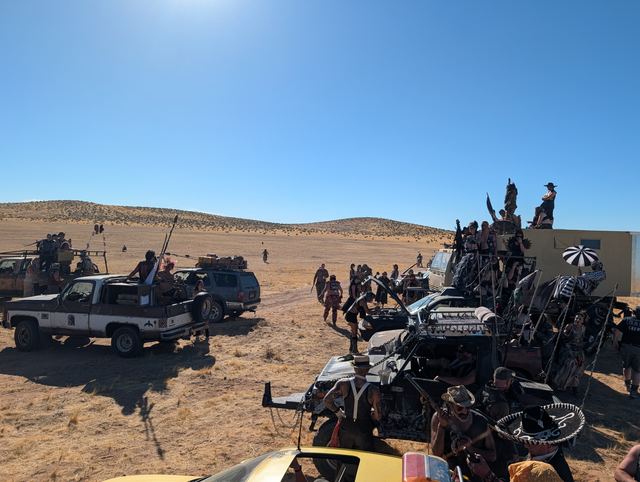 The convoy viewed from atop a vehicle, riders waving weapons from the roofs.
