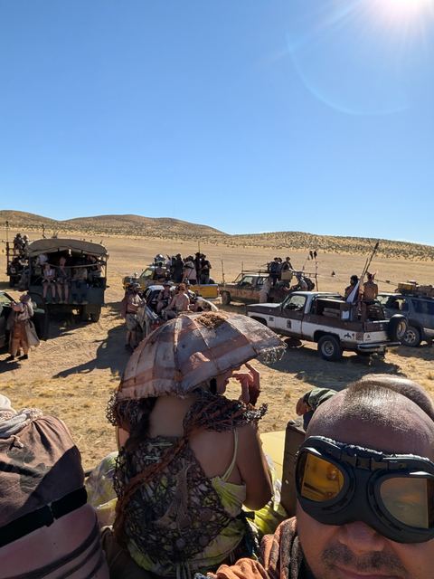 Two riders in goggles and a woven hat on a vehicle roof, convoy below.
