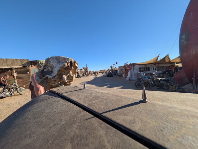 The skull hood ornament seen from behind as the war car drives past shacks.