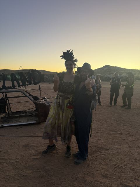 A woman in a feathered headdress and a man in a hat at dusk in the desert.