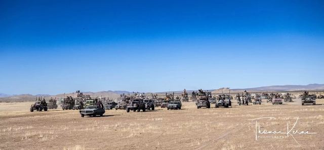 Distant convoy vehicles spread across the dry flats under blue sky. Photographer's watermark in the corner.