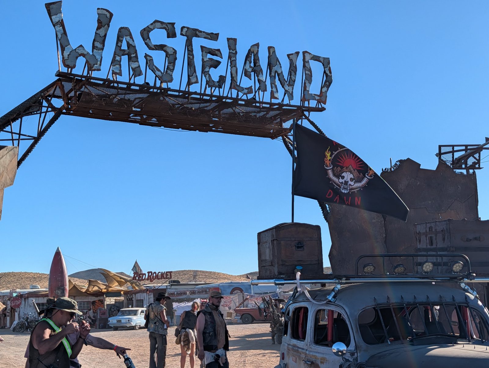 The tribe's black flag with its red emblem flying from the war car beneath the WASTELAND gate sign.