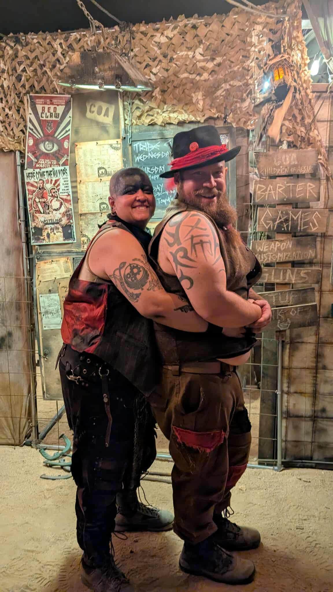 Two members hug for the camera in front of a barter-town stall, chalkboards listing games and trade rules behind.