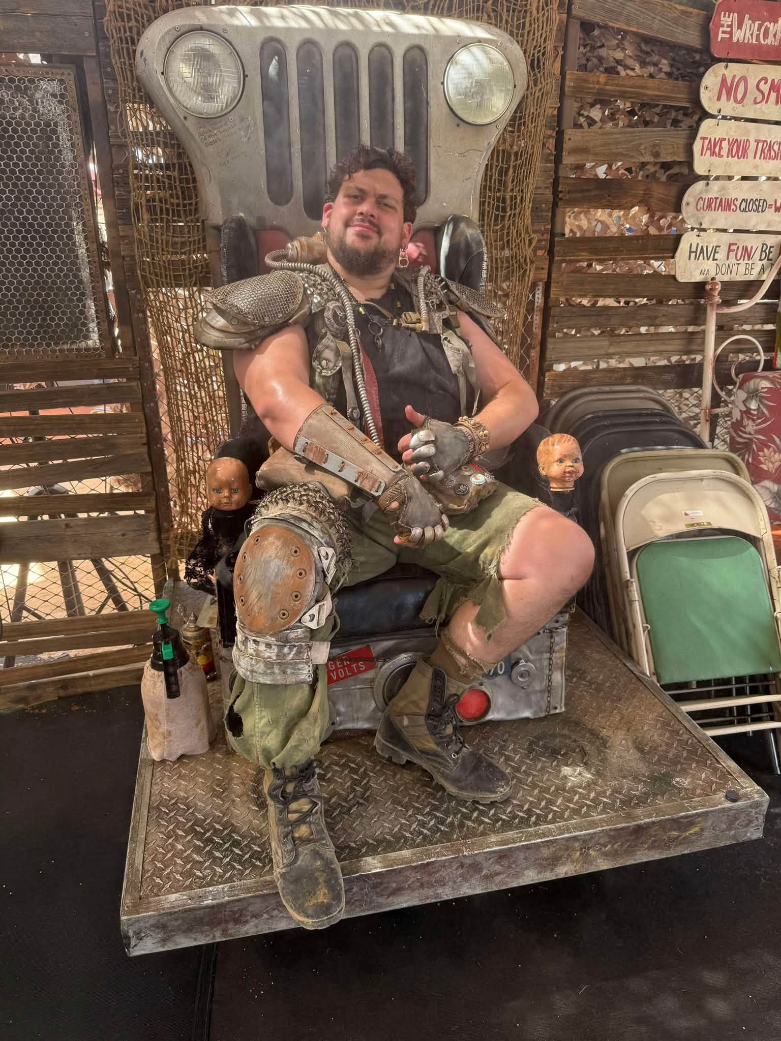 Brisket in spiked pauldron and rusted armor plates lounges on a throne built from a Jeep front grille, baby-doll heads mounted on either side.