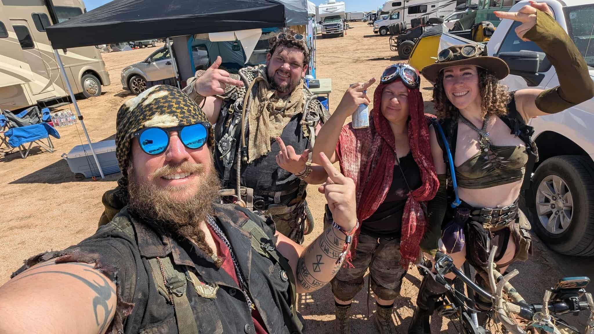 Daytime group selfie at camp — four wastelanders in distressed gear, goggles, and headwraps grinning under a blue desert sky, canopies and vehicles behind.