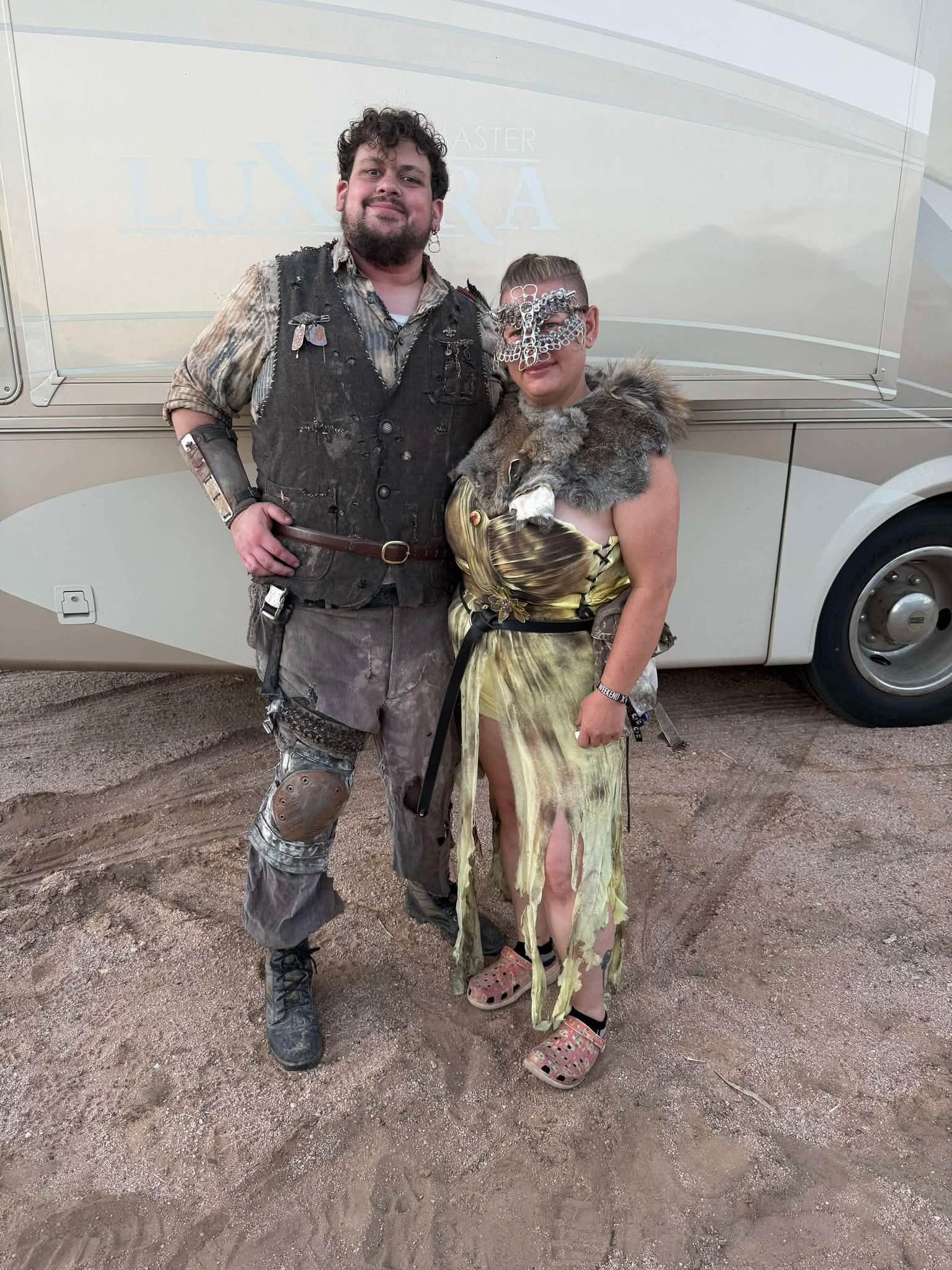 Champ Chicharrón in a torn gold dress, fur mantle, and chainmail face mask stands beside Brisket in a distressed vest, in front of an RV.