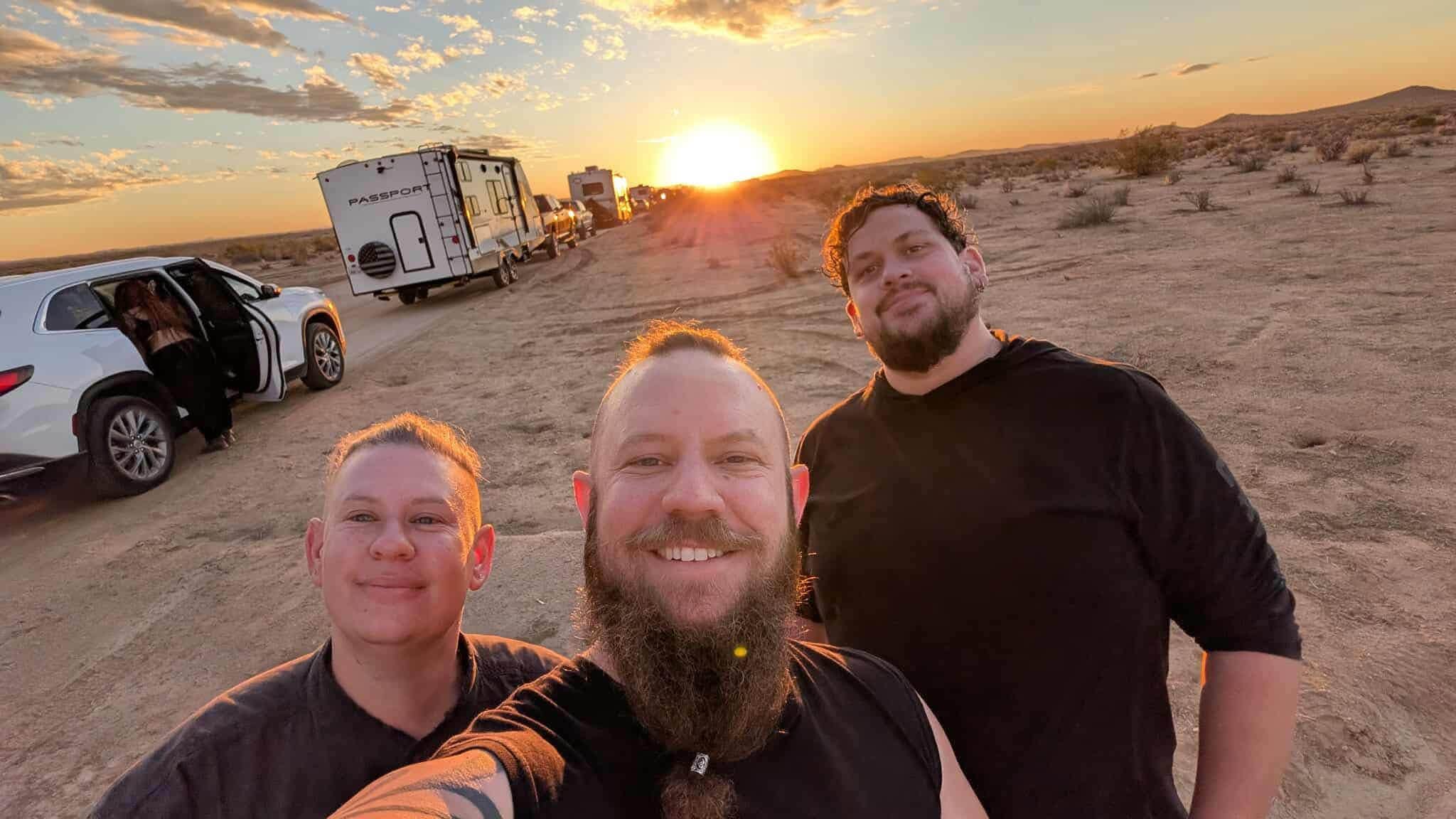 Three tribe members take a selfie on a dirt road at sunset, the sun blazing low between trailers and RVs lined up behind them.