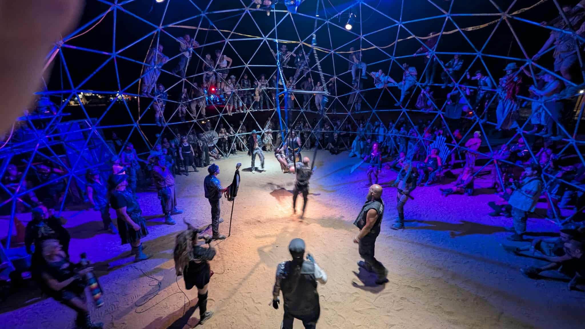 The geodesic thunderdome at night washed in blue light, spectators clinging to the struts above the sand floor.