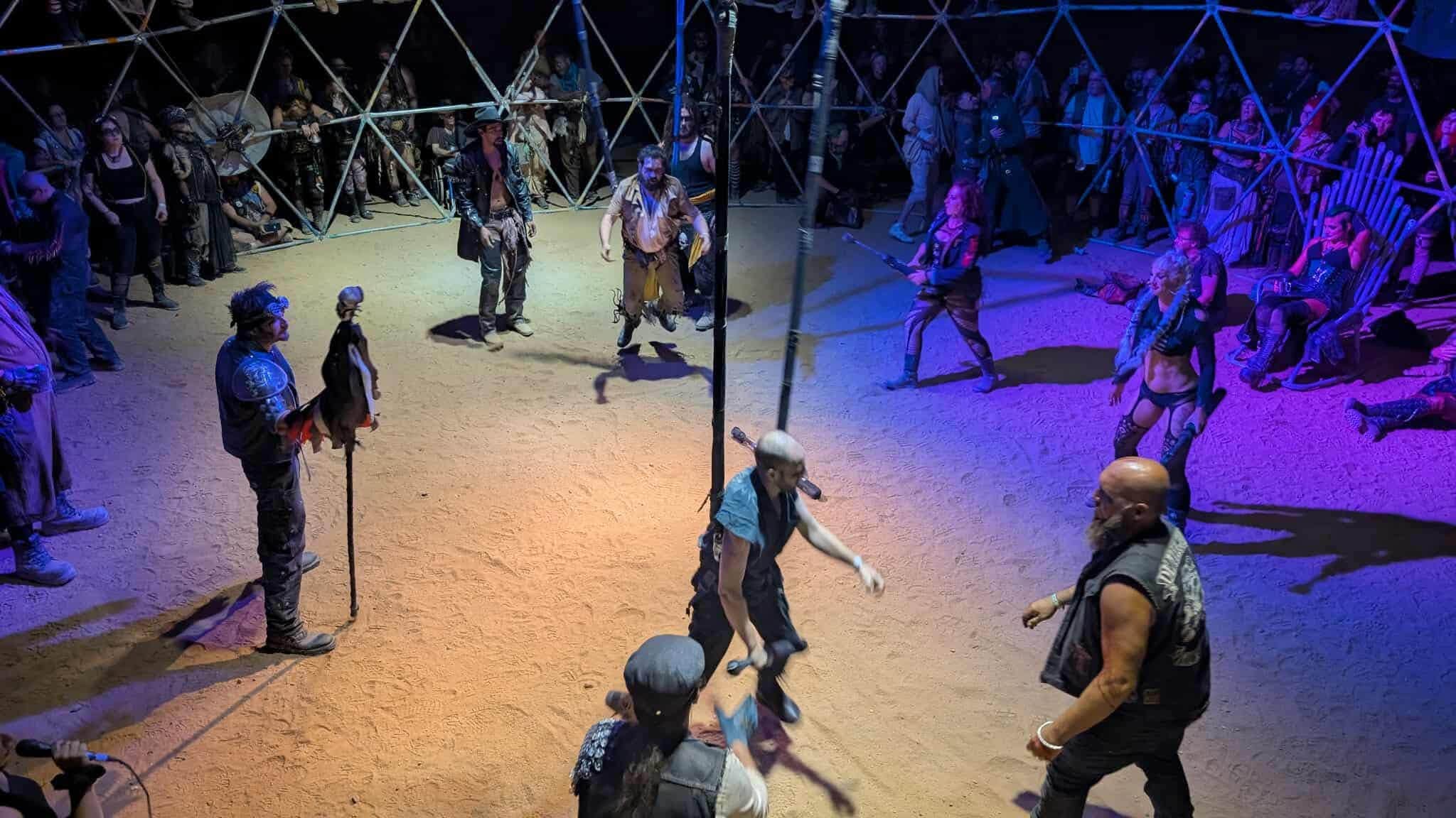 Fighters square off on the sand inside the thunderdome while a referee with a feathered staff watches, the crowd pressed against the lattice.