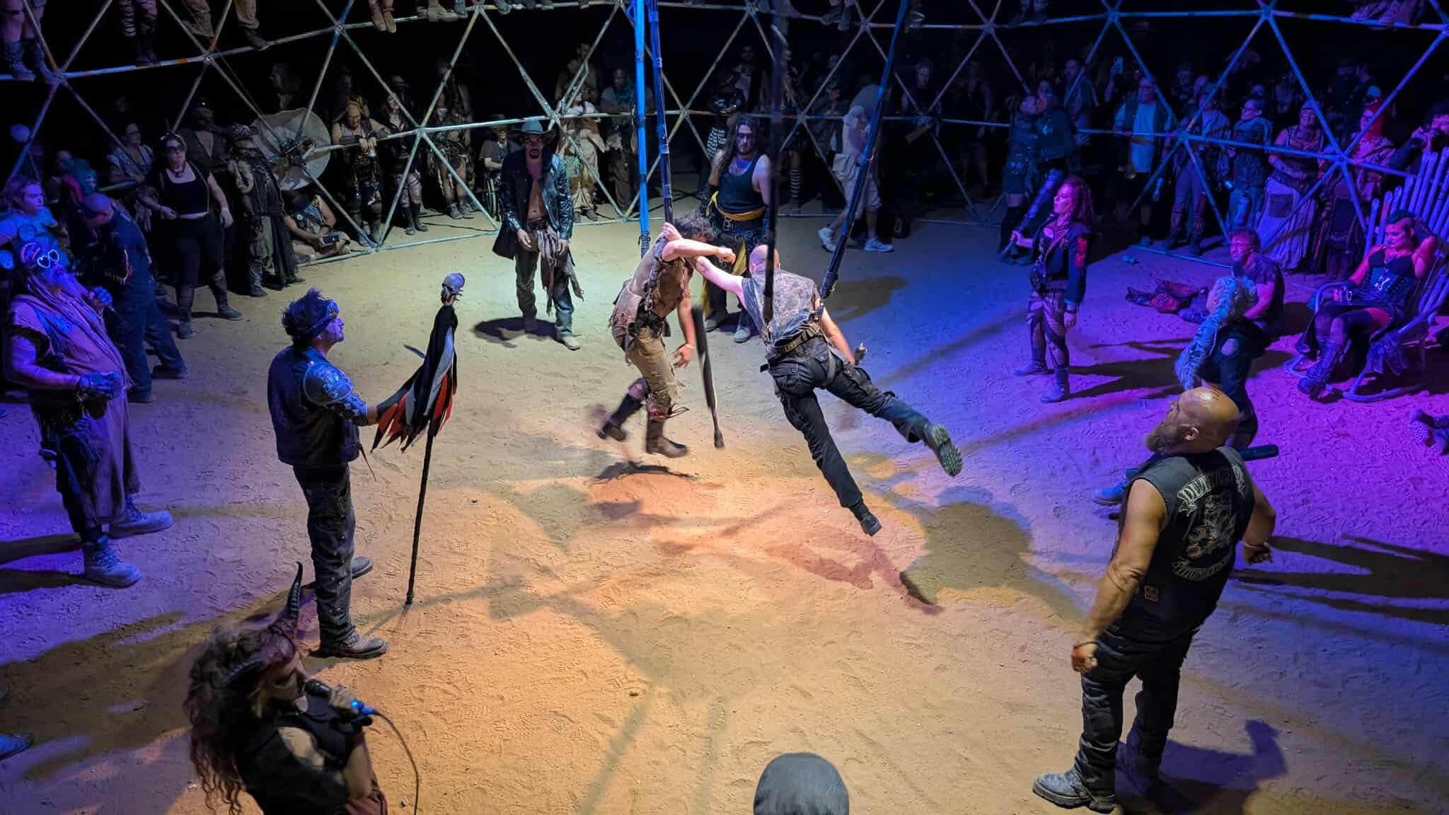 A fighter leaps mid-swing inside the thunderdome as the crowd watches from the lattice and the sand.