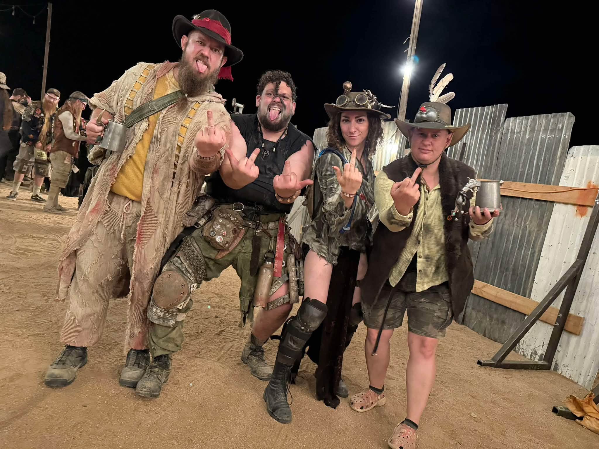 Four tribe members in full kit at night — tin mugs in hand, tongues out, every middle finger raised at the camera.