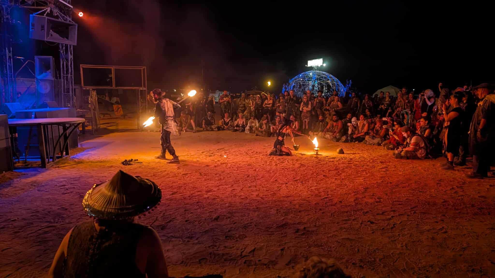 A fire performer spins twin flames at the center of a torch-lit night crowd, stage rigging glowing at the left.