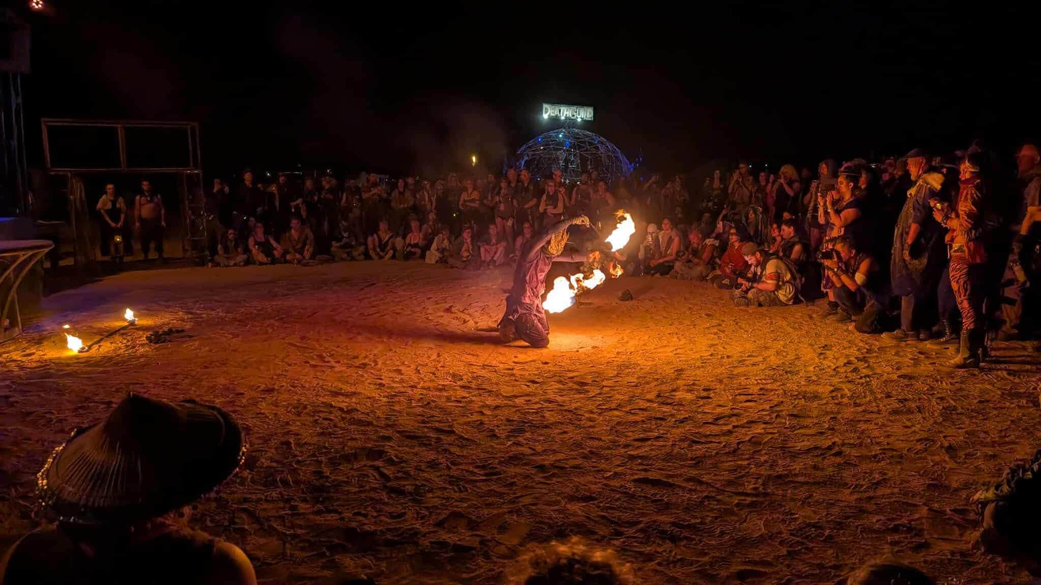 A fire performer kneels in the sand, arms spread, flames blooming from both hands before a night crowd.