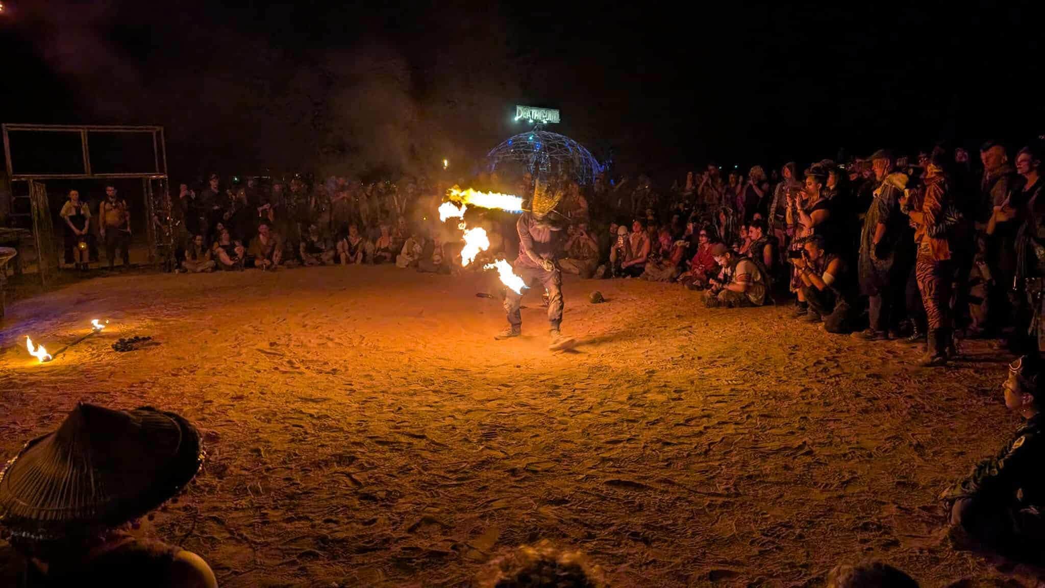 A fire performer in a horned mask breathes a jet of flame before a night crowd ringed around a sand arena, a geodesic dome glowing behind.