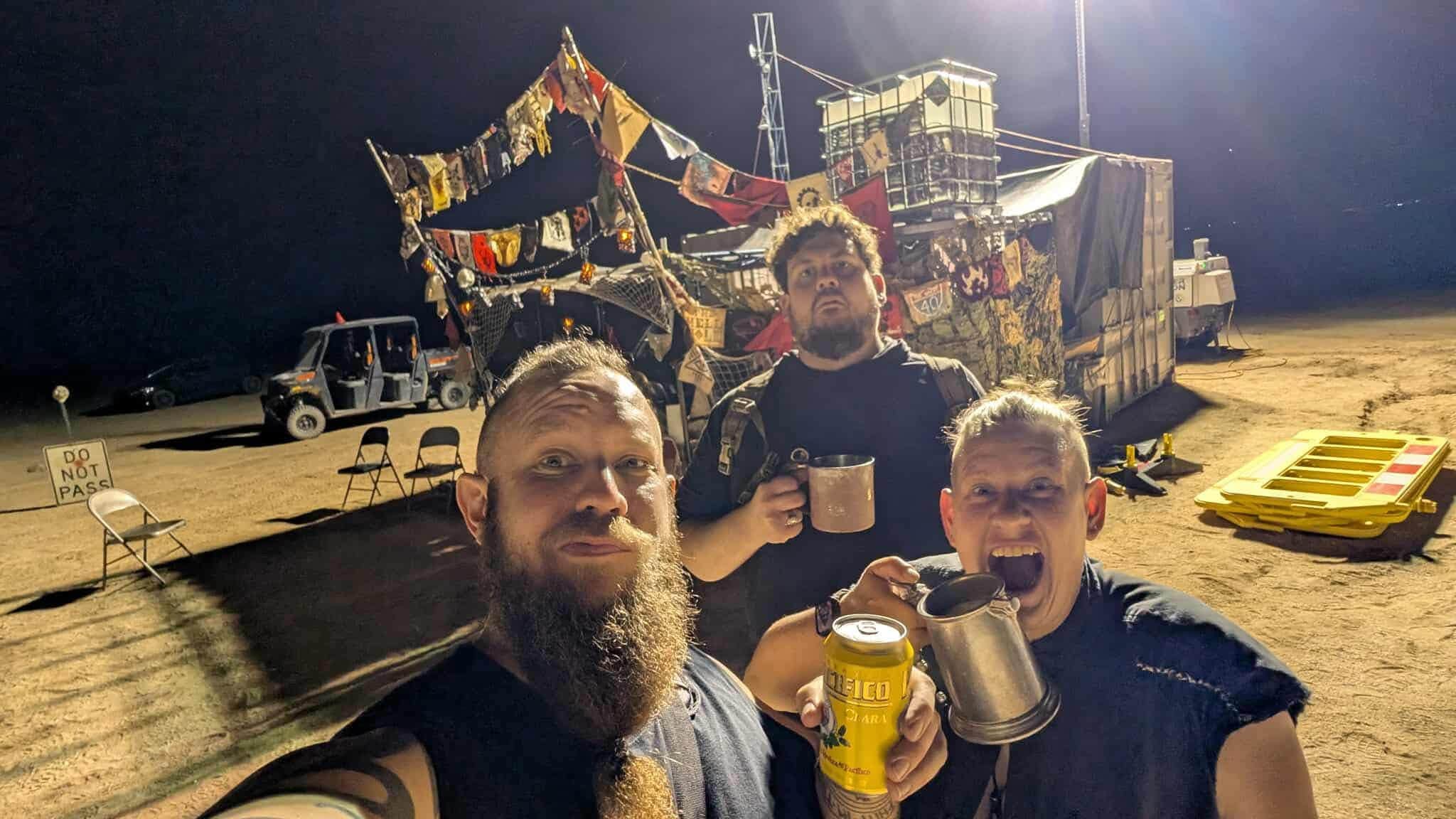 Three members raise metal mugs and a can at night in front of a camp frontage strung with banners, patches, and a DO NOT PASS sign.