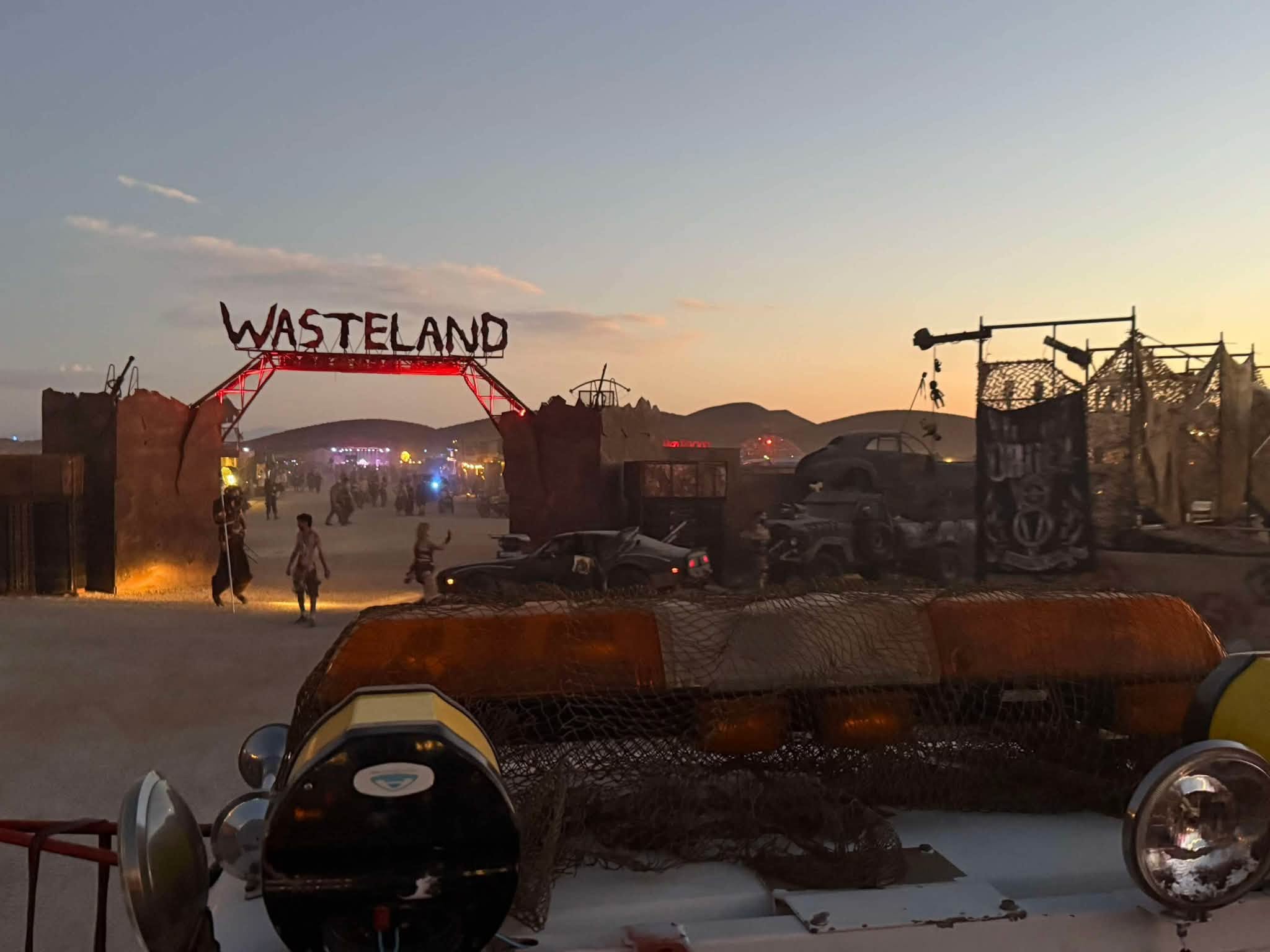 The WASTELAND gate at dusk with wrecked-car set pieces and quads parked along the scrap walls.