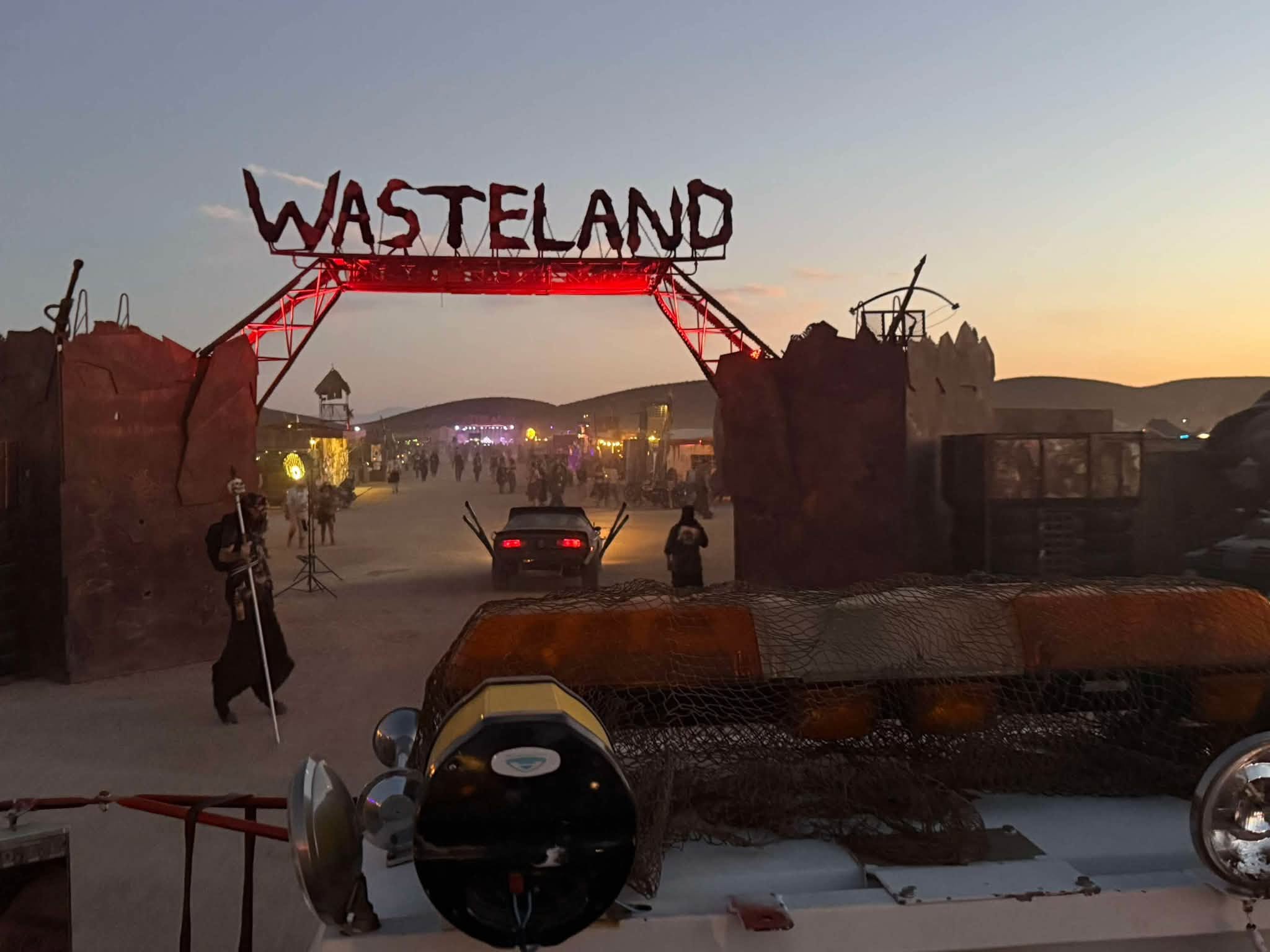 The Wasteland Weekend gate at dusk — a metal arch spelling WASTELAND in red over scrap-walled towers, seen from atop a vehicle, with wastelanders walking the dusty road below.