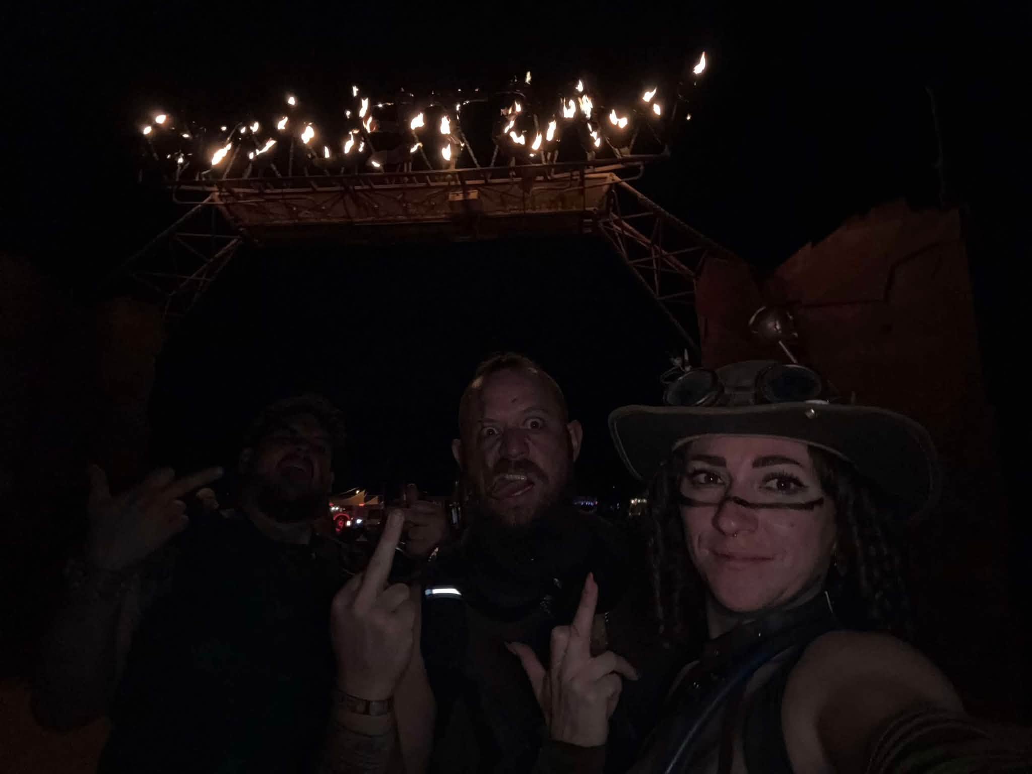 Three members at night beneath the gate, the WASTELAND letters lit by open flame high above.