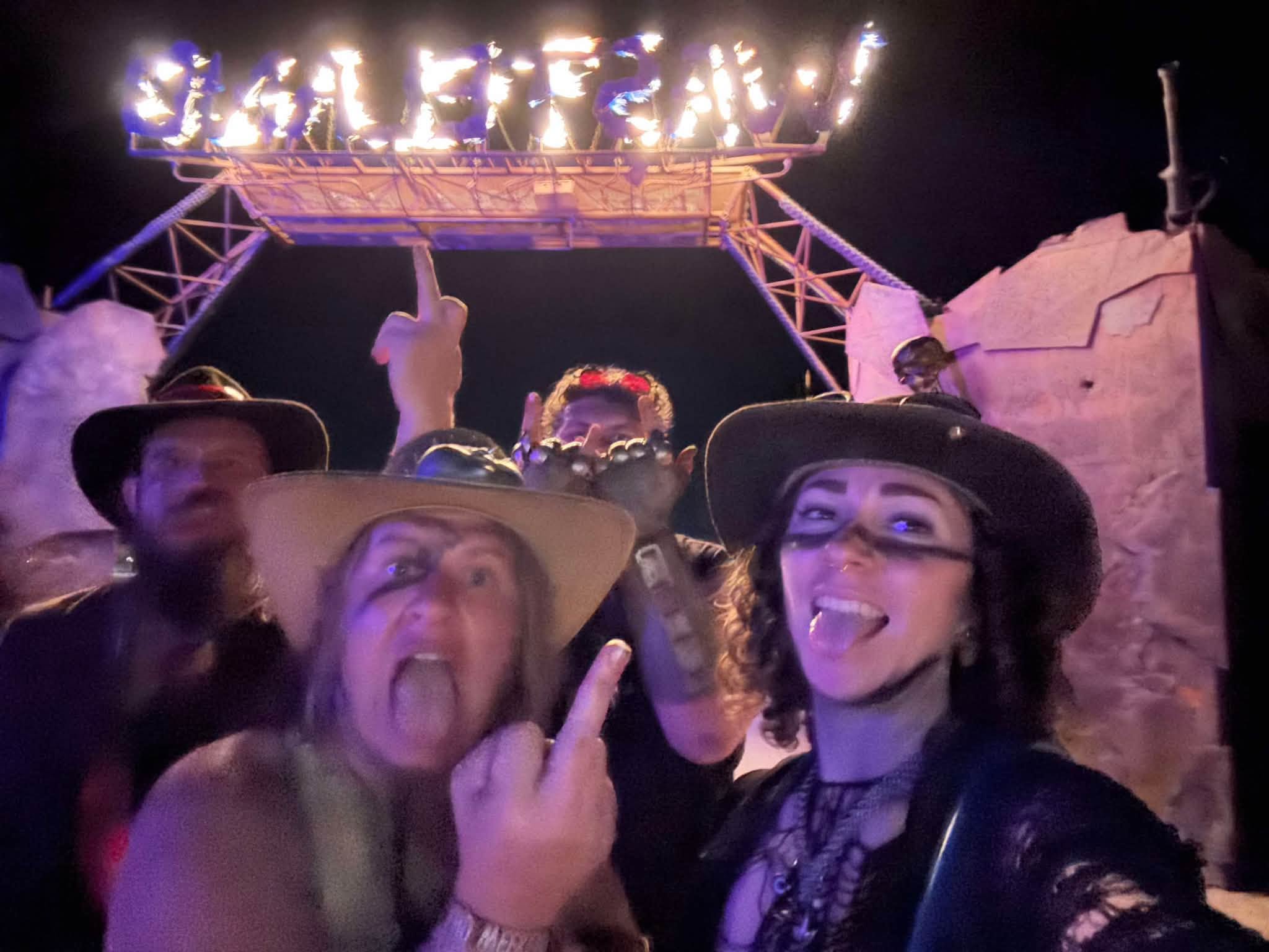 Night selfie under the gate, the WASTELAND letters burning with open flame overhead while five tribe members mug for the camera.
