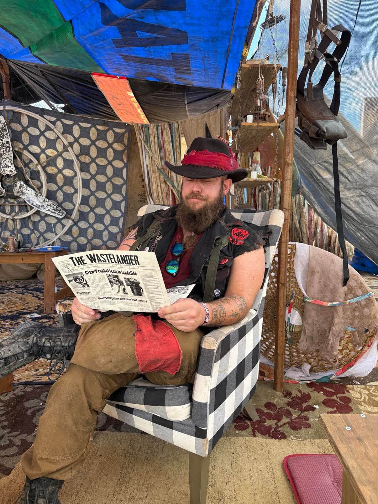 Grandpa in a red-banded black hat reads The Wastelander newspaper in a plaid armchair inside a draped trading post — the exact scene on his badge.
