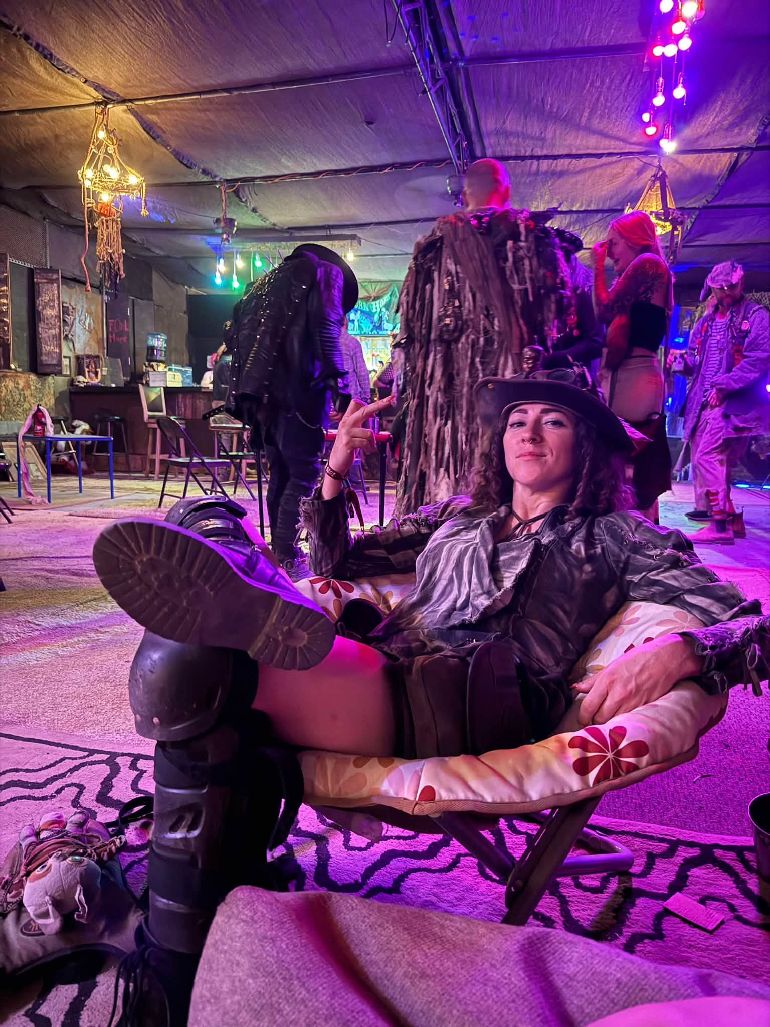 A member in a brimmed hat lounges sideways across a camp chair in a purple-lit tent, boots over the armrest.