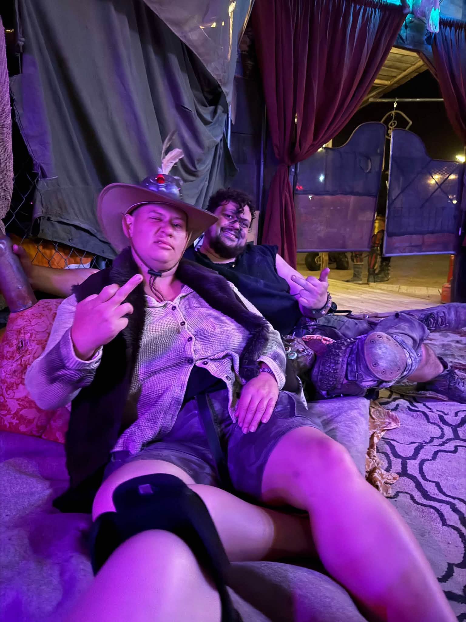 Two members sprawl across cushions in a purple-lit lounge tent, flashing hand signs at the camera.