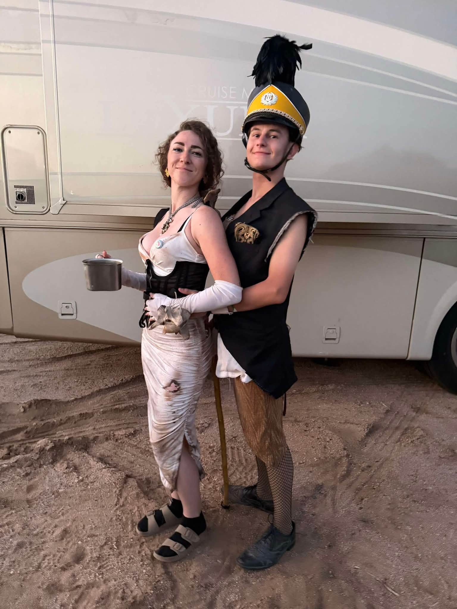 Two wastelanders pose back to back in front of an RV — one in a torn white dress holding a tin cup, one in a feathered marching-band shako and cut-off blazer.