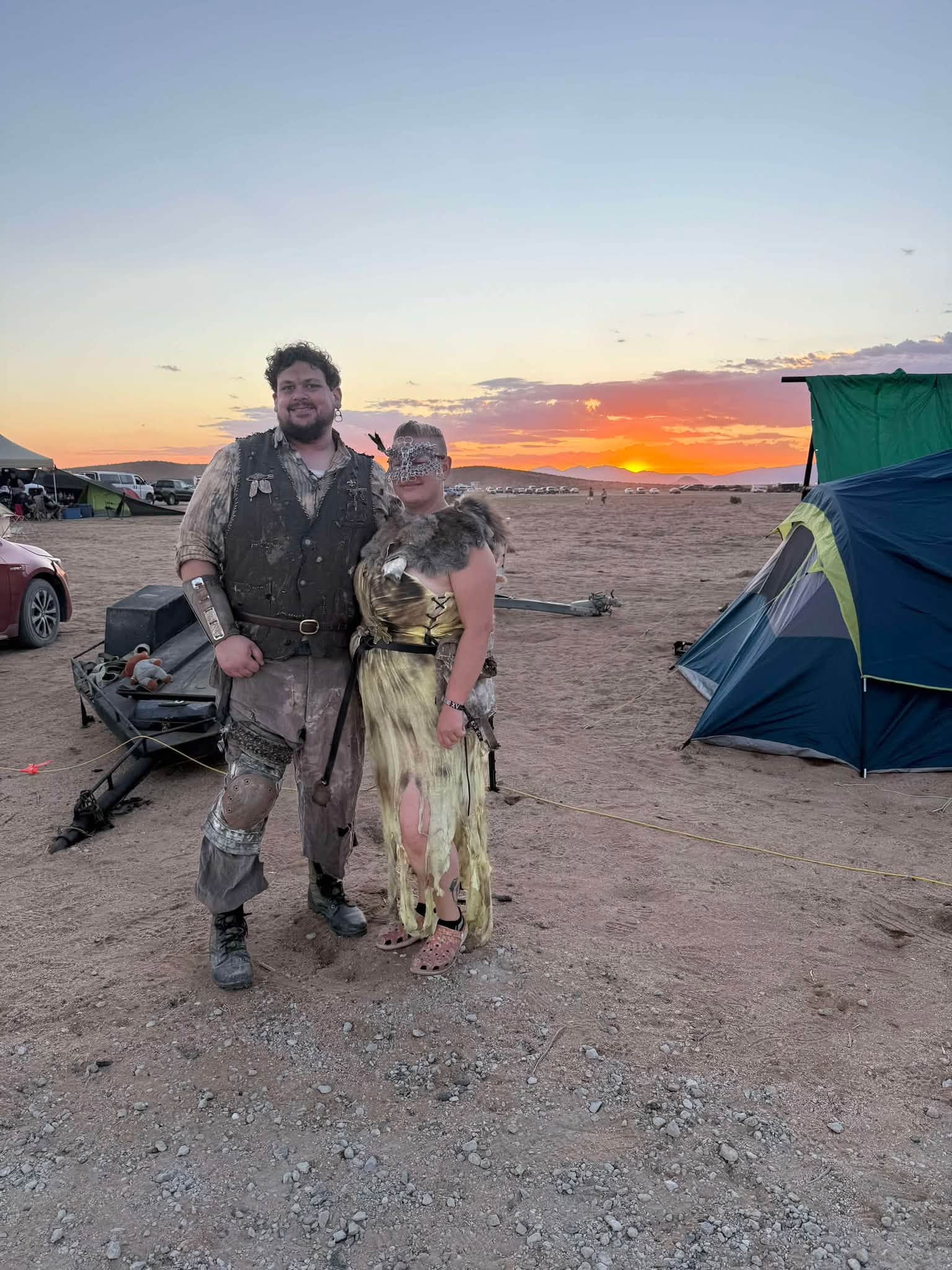 Two members in full kit — distressed vest and torn gold dress with a fur mantle — pose at golden hour, the sun setting between camp tents behind them.