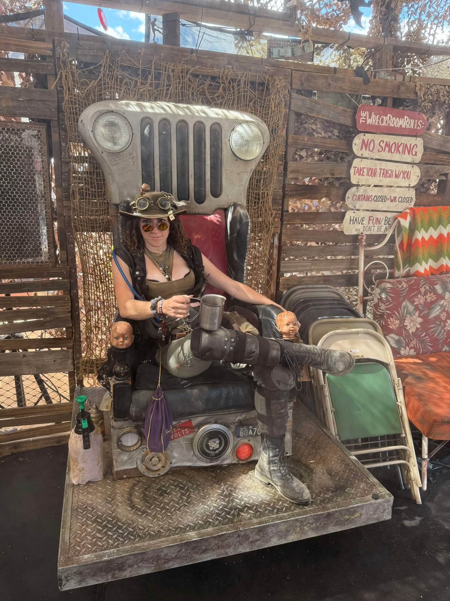 A wastelander in a brimmed hat and goggles takes the Jeep-grille throne, boot up on the deck plate, doll heads on the armrests.