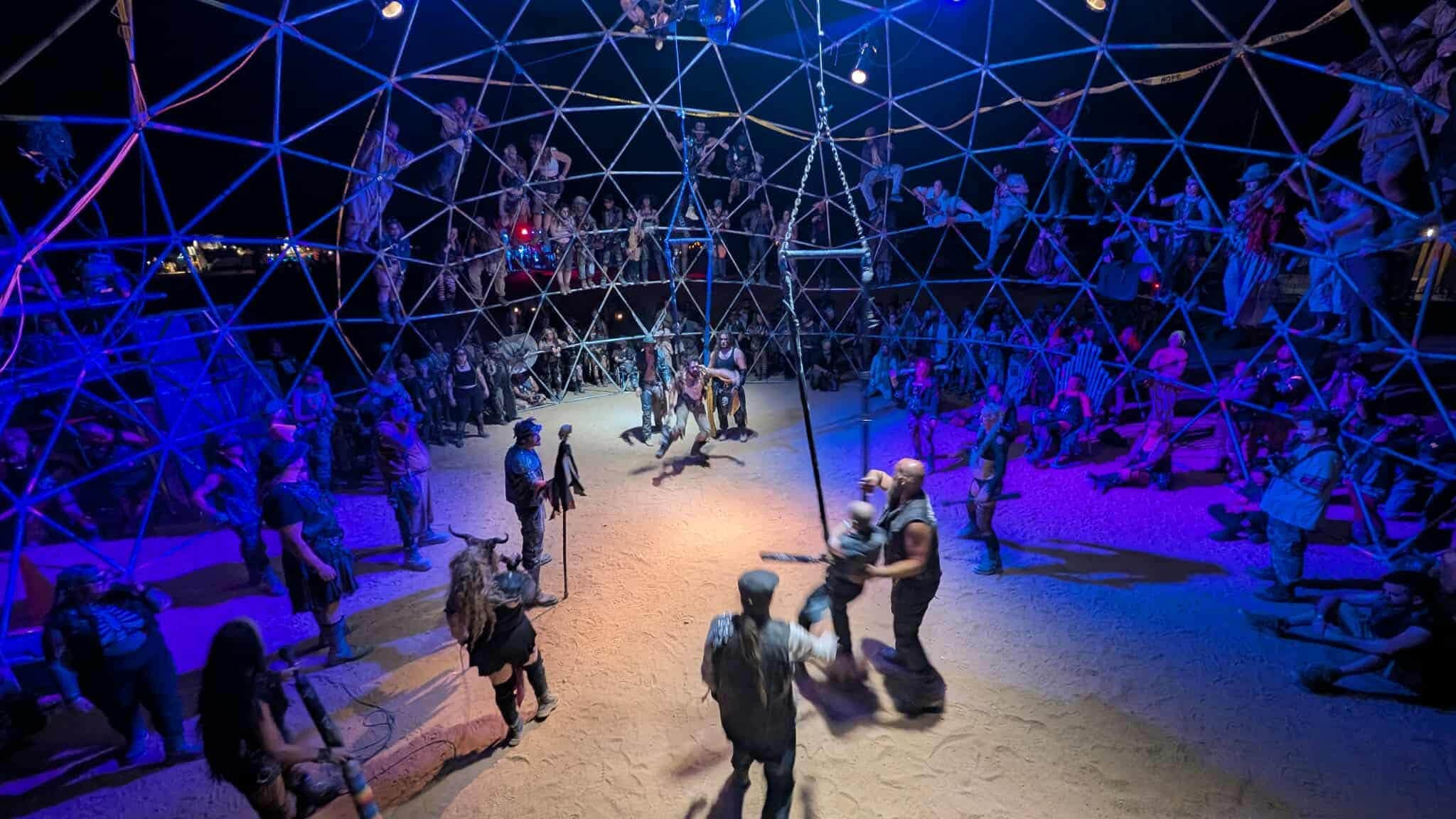 Inside a geodesic dome at night lit blue and orange, spectators climb the struts while fighters square off on the sand floor below.