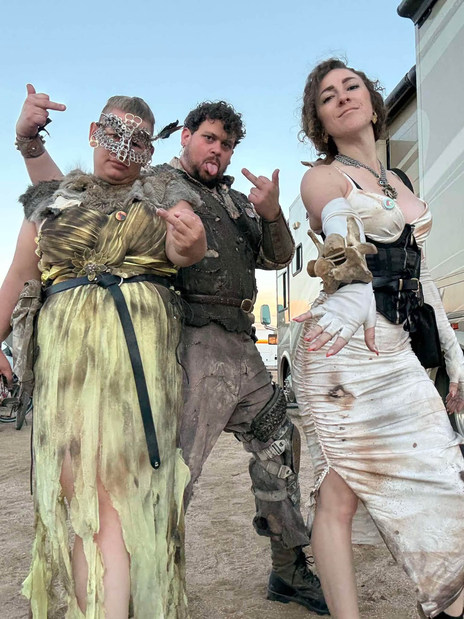 Three members at dusk by the RVs — chainmail mask and gold dress, tongue out in distressed leathers, and a torn white dress with a bone bracer.