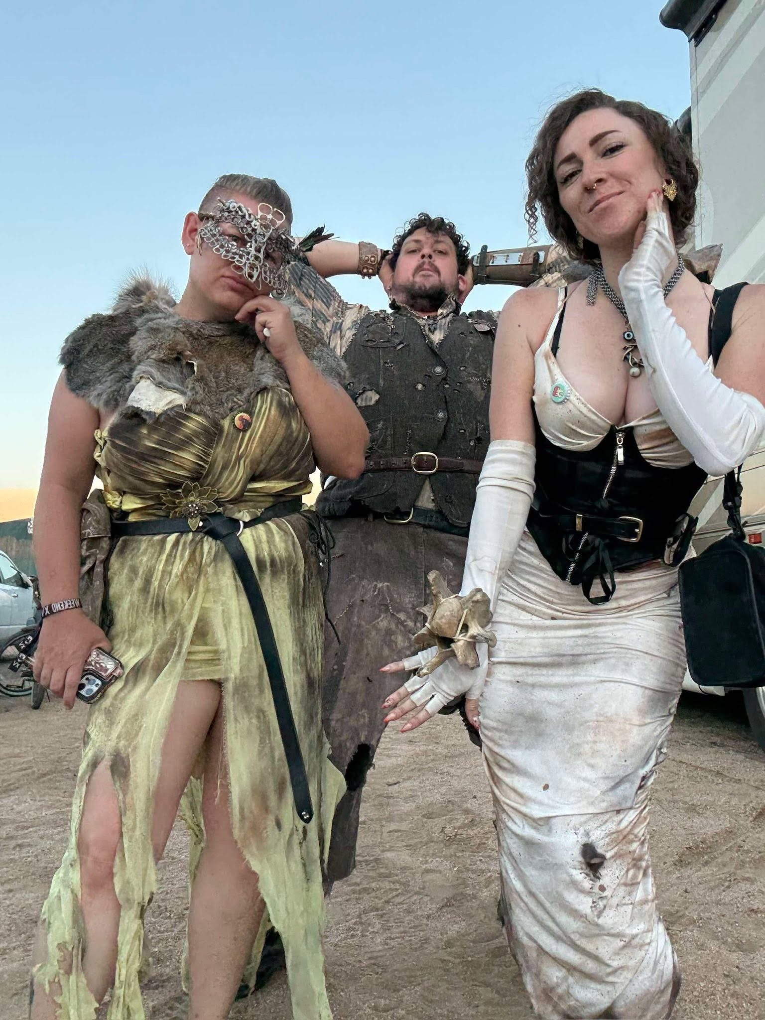 Three members pose beside an RV at dusk, one peering over the others' shoulders from behind.