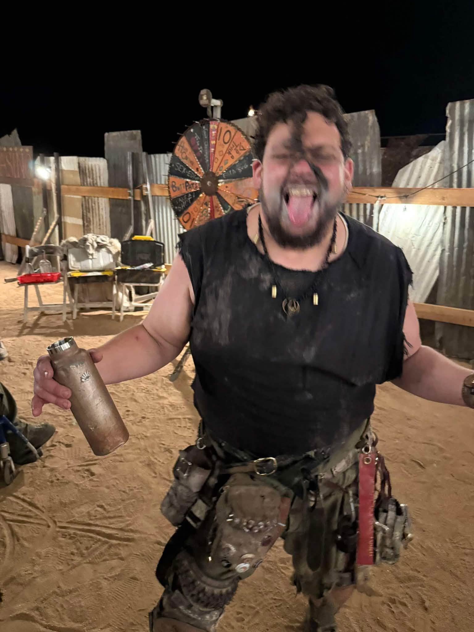 Brisket at night, tongue out and arms spread, holding a battered steel bottle, a barter wheel of fortune behind him.